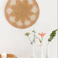 Woven wall art piece on a white wall with decorative vases and candles on a wooden surface.