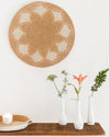 Woven wall art piece on a white wall with decorative vases and candles on a wooden surface.